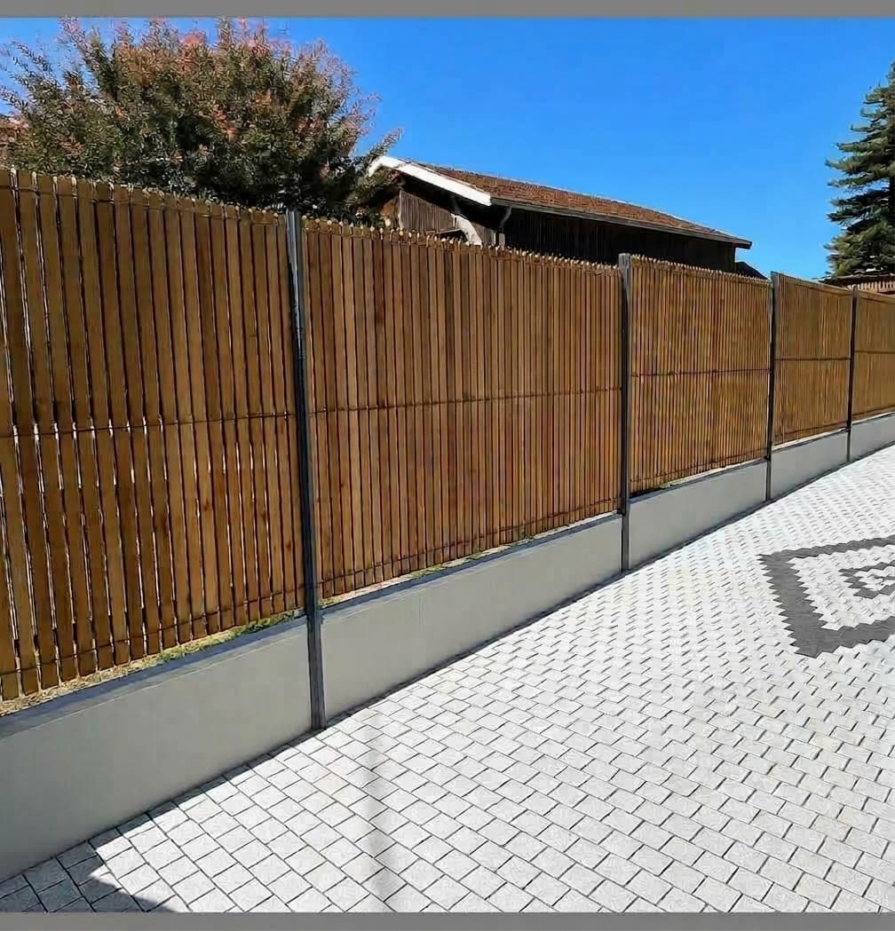 Clôture en lattes de bois sur muret en béton longeant une allée pavée.